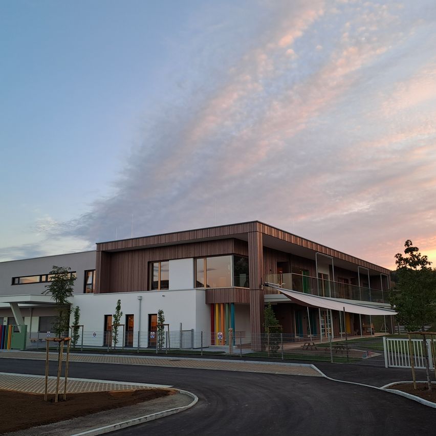 Ein modernes Gebäude mit brauner Fassade und Glasfenstern ist unter einem bewölkten Himmel zu sehen. Bäume und Pflanzen sind in der Nähe.