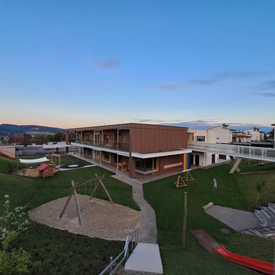 Luftaufnahme eines modernen Gebäudes mit Spielplatz. Es hat einen Rasen mit Schaukeln, Rutschen und einer Regenbogenwand. Das Gebäude hat Glasfenster und eine Brücke.