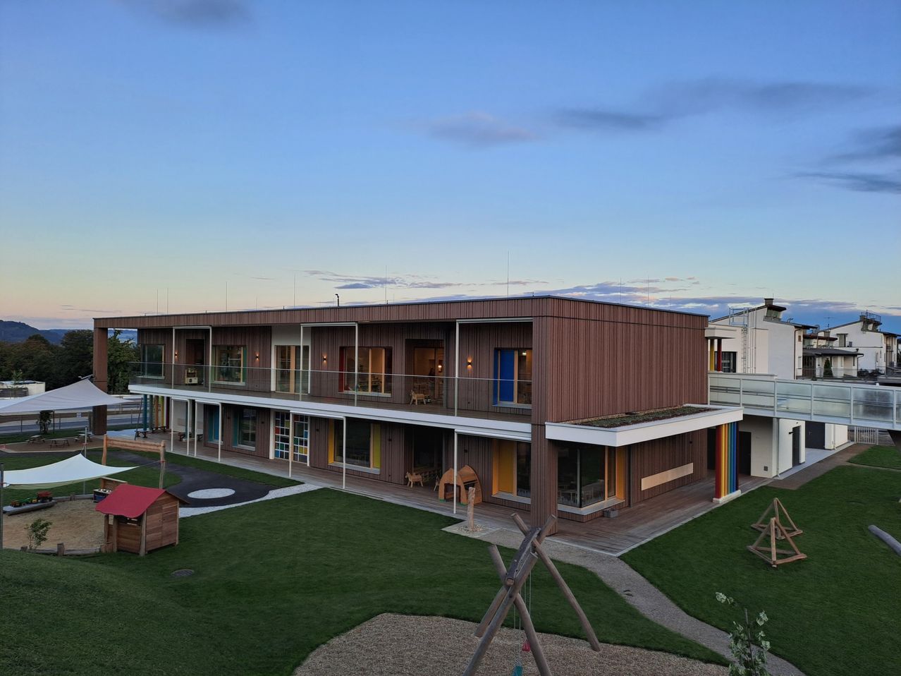 Ein modernes Gebäude mit mehreren Etagen und einem grünen Dach, umgeben von einem gepflegten Rasen und einem Spielbereich.