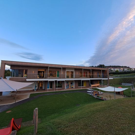 Ein Blick aus der Luft auf ein modernes Gebäude mit mehreren Balkonen und einem Spielplatz davor. Das Gebäude ist von einem Rasenfeld umgeben und hat einen klaren blauen Himmel.