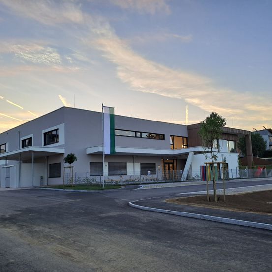 Das Bild zeigt ein modernes Gebäude mit einer Flagge davor. Das Gebäude hat eine graue Außenseite mit weißen Fenstern und einem gebogenen Eingang. Ein Baum ist in der Nähe des Eingangs gepflanzt. Der Himmel ist blau mit einigen Wolken.