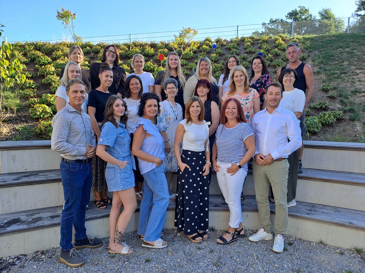 Eine Gruppe von Männern und Frauen steht auf Betonstufen und posiert für ein Foto. Sie sind in verschiedenen Outfits gekleidet und einige tragen Brillen. Der Hintergrund zeigt einen Hügel mit Pflanzen und einem Zaun.
