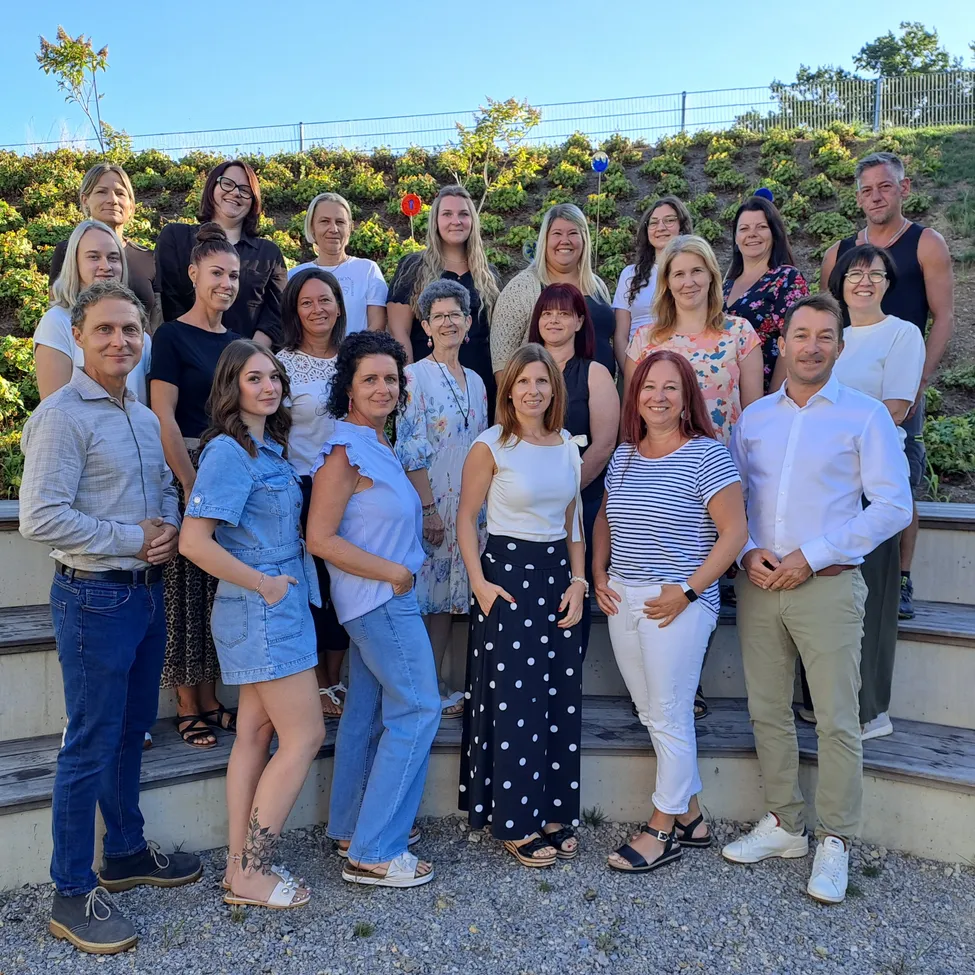 Eine Gruppe von Männern und Frauen steht auf Betonstufen und posiert für ein Foto. Sie sind in verschiedenen Outfits gekleidet und einige tragen Brillen. Der Hintergrund zeigt einen Hügel mit Pflanzen und einem Zaun.