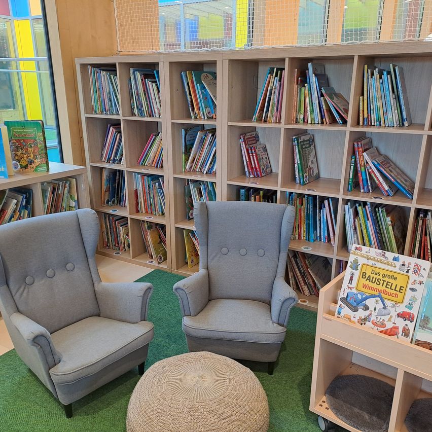 Eine gemütliche Leseecke mit zwei grauen Sesseln und einem runden Pouf. Dahinter befinden sich Bücherregale mit Kinderbüchern. Ein grüner Teppich verleiht Wärme.