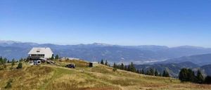 Ein landschaftlicher Blick von einem Berggipfel zeigt ein kleines Gebäude, ein geparktes Auto und eine Person, die auf einem Pfad geht. Die Landschaft ist umgeben von Bäumen und Bergen unter einem klaren blauen Himmel.