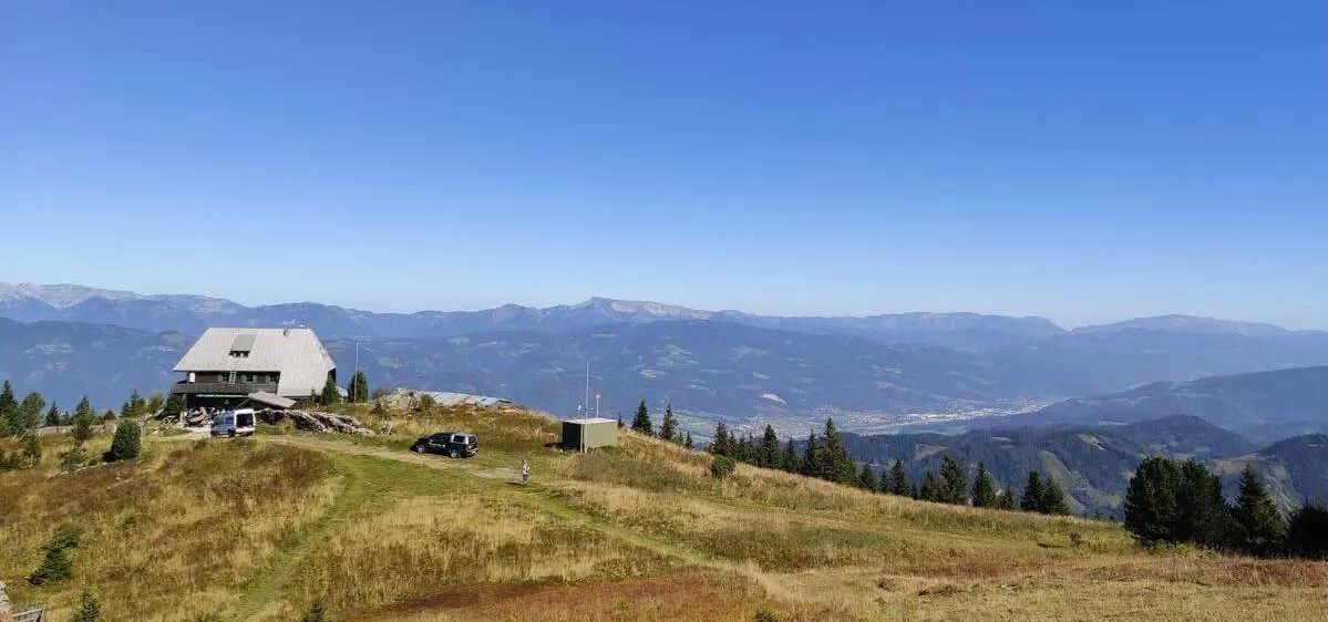 Ein landschaftlicher Blick von einem Berggipfel zeigt ein kleines Gebäude, ein geparktes Auto und eine Person, die auf einem Pfad geht. Die Landschaft ist umgeben von Bäumen und Bergen unter einem klaren blauen Himmel.