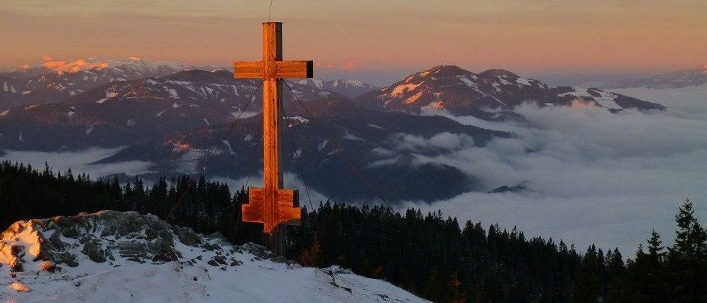Ein hölzerne Kreuz steht auf einem verschneiten Berggipfel, mit einem Kiefernwald darunter und schneebedeckten Bergen in der Ferne.