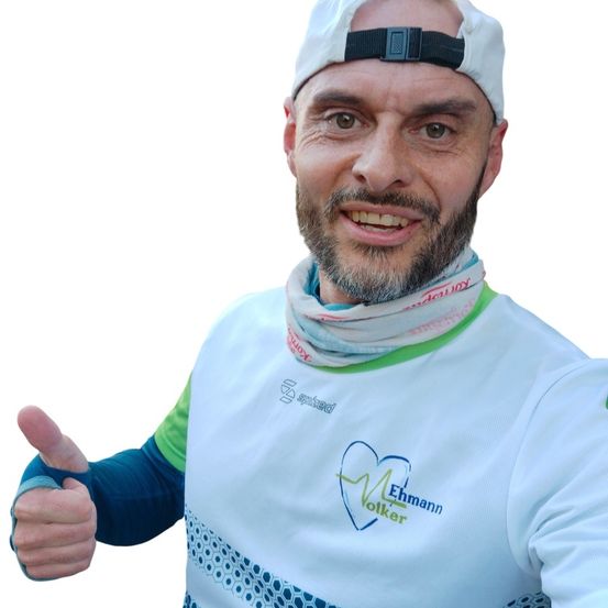 Ein Mann in einem weißen Shirt mit grünem und blauem Design lächelt und gibt einen Daumen hoch. Er trägt eine weiße Kappe mit einem schwarzen Riemen.