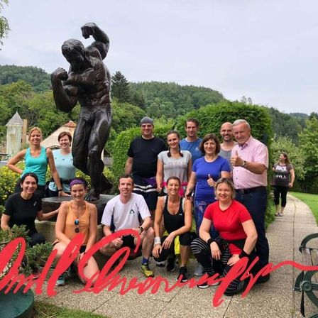 Eine Gruppe von Menschen in Sportkleidung posiert für ein Foto vor einer muskulösen Statue, mit einer malerischen Bergkulisse im Hintergrund.