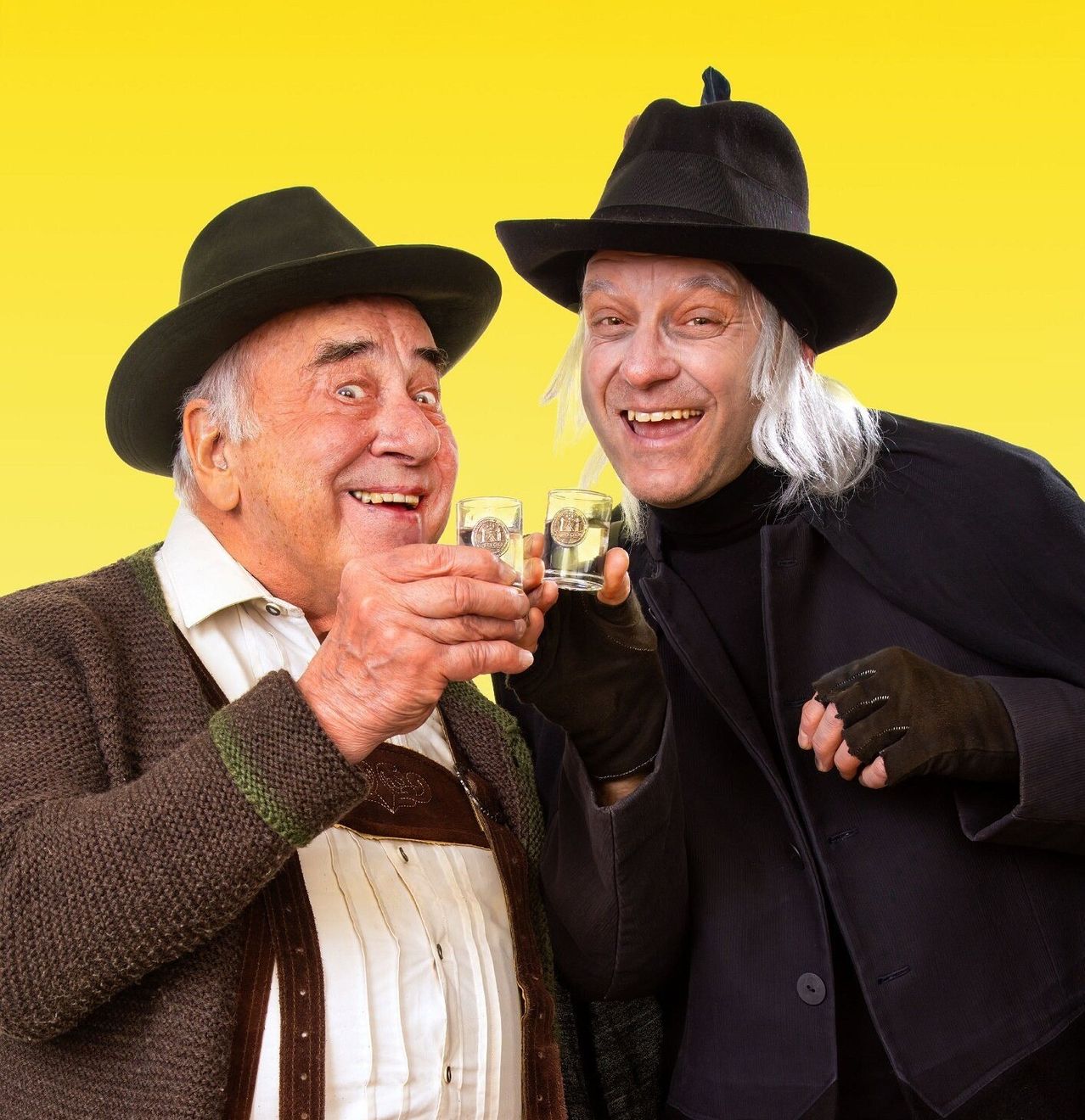 Zwei Männer, einer in einem gestrickten Pullover und Hut, der andere in einem schwarzen Mantel, halten kleine Gläser und lächeln für die Kamera vor gelbem Hintergrund.