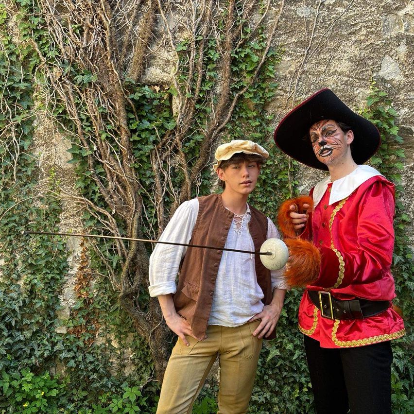 Zwei junge Männer in historischen Kostümen posieren mit Schwertern in einem Garten. Einer trägt ein Löwenkostüm und der andere einen Piratenhut.