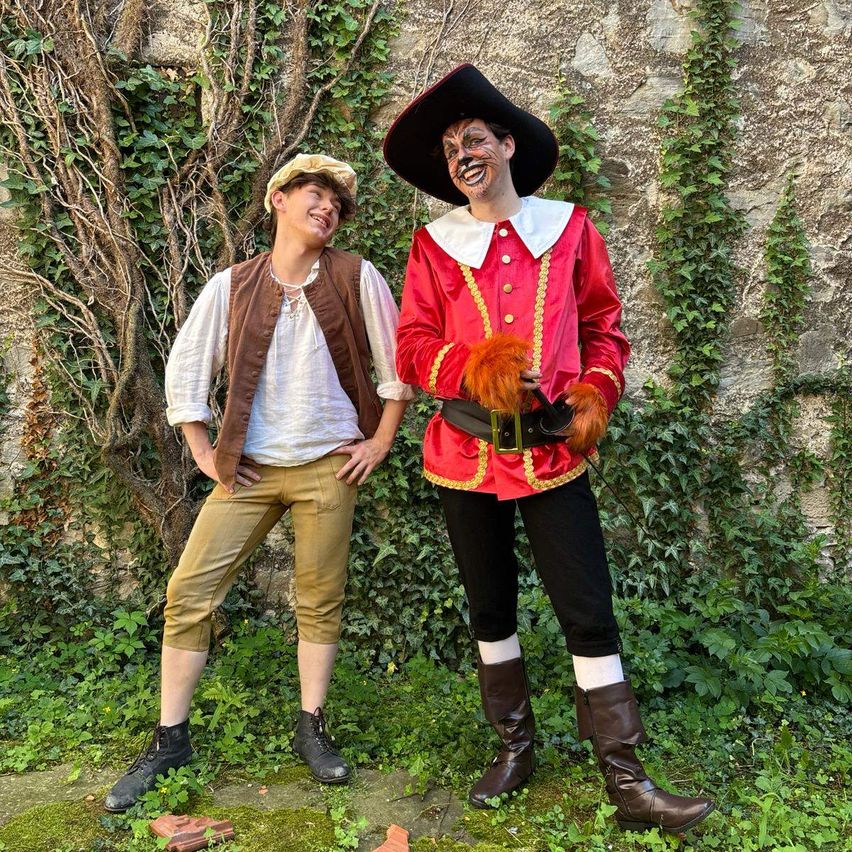 Zwei Personen in historischen Kostümen, eine in einem roten und schwarzen Ensemble mit Hut und die andere in einem weißen Hemd und einer braunen Weste, stehen nebeneinander in einem Garten.