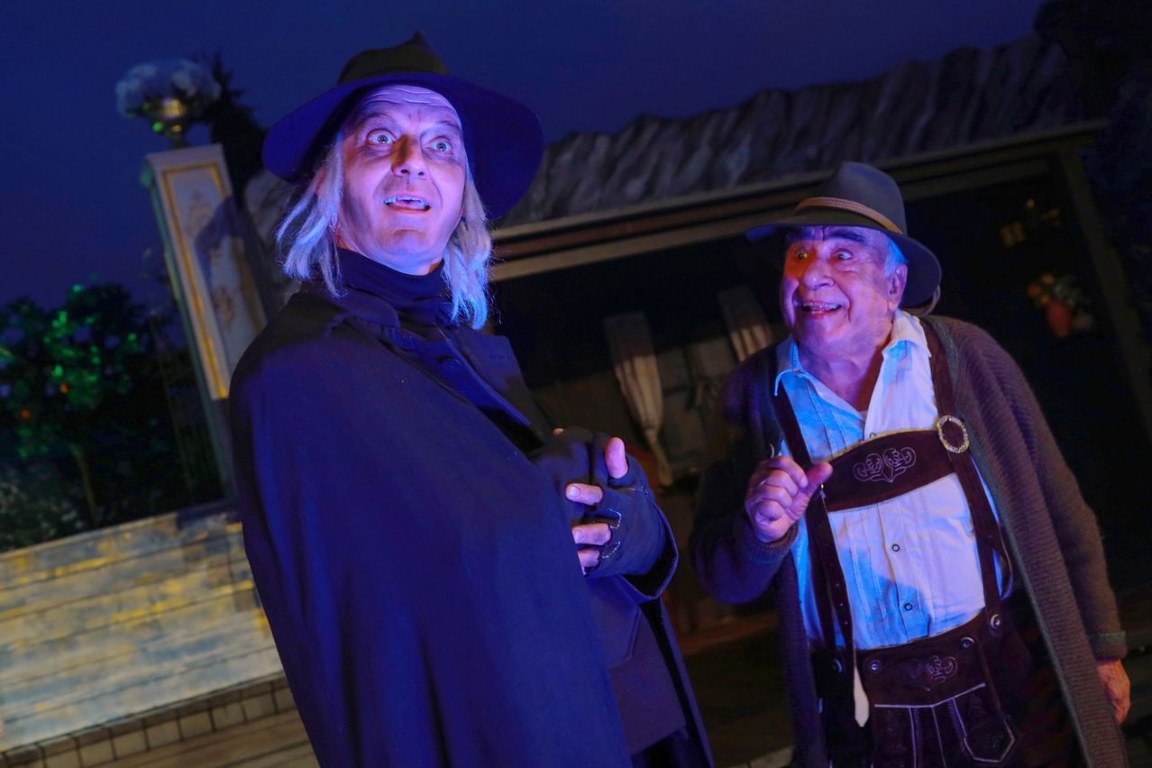 Two men in costume, one in a blue cloak and hat, the other in a white shirt and suspenders, stand next to each other in a dimly lit outdoor setting.