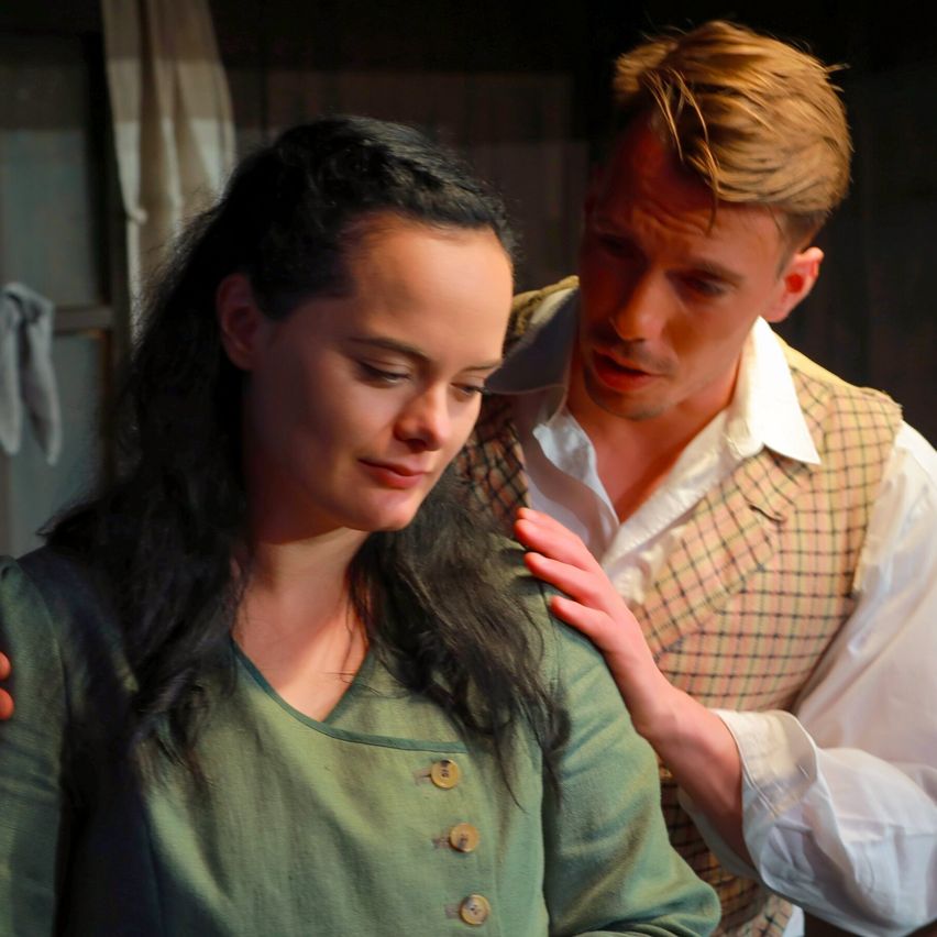 A woman with long dark hair and a green dress looks down while a man with a plaid vest touches her shoulder.