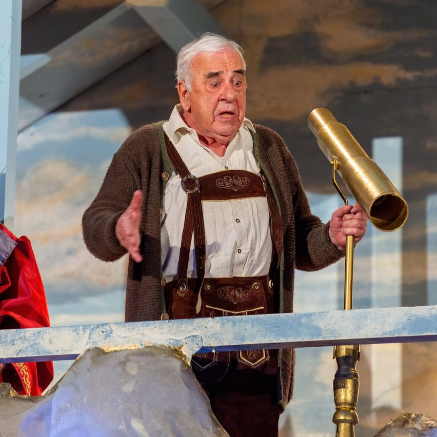 An elderly man in traditional Bavarian attire, holding a golden telescope, stands on a stage, possibly performing in a play.