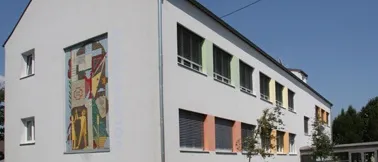 Ein modernes weißes Gebäude mit farbenfrohen Fenstern und einer großen Wandmalerei an der Seite, unter einem klaren blauen Himmel.