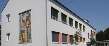 Ein modernes weißes Gebäude mit farbenfrohen Fenstern und einer großen Wandmalerei an der Seite, unter einem klaren blauen Himmel.