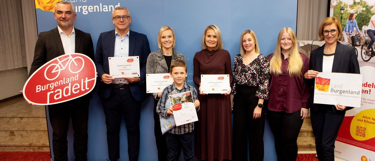 Eine Gruppe von Menschen posiert für ein Foto vor einem blauen Hintergrund. Jeder hält ein Zertifikat in der Hand, und ein Junge hält ein Buch. Sie lächeln und scheinen zu feiern.