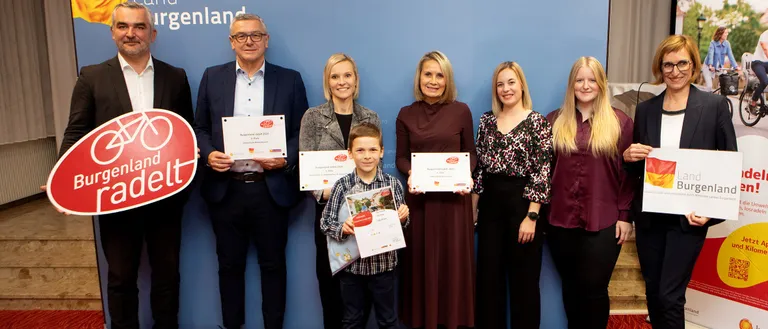 Eine Gruppe von Menschen posiert für ein Foto vor einem blauen Hintergrund. Jeder hält ein Zertifikat in der Hand, und ein Junge hält ein Buch. Sie lächeln und scheinen zu feiern.