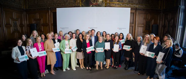 Eine Gruppe von Frauen und Männern in formeller Kleidung posiert mit Zertifikaten in der Hand für ein Foto. Sie stehen vor einem großen weißen Banner mit Logos und Text. Ein Gemälde einer historischen Figur ist hinter ihnen.