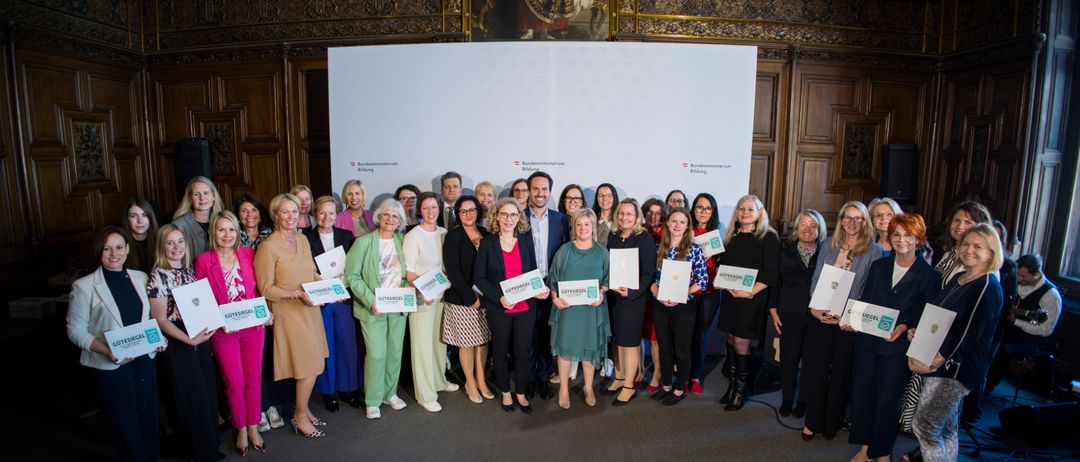 Eine Gruppe von Frauen und Männern in formeller Kleidung posiert mit Zertifikaten in der Hand für ein Foto. Sie stehen vor einem großen weißen Banner mit Logos und Text. Ein Gemälde einer historischen Figur ist hinter ihnen.