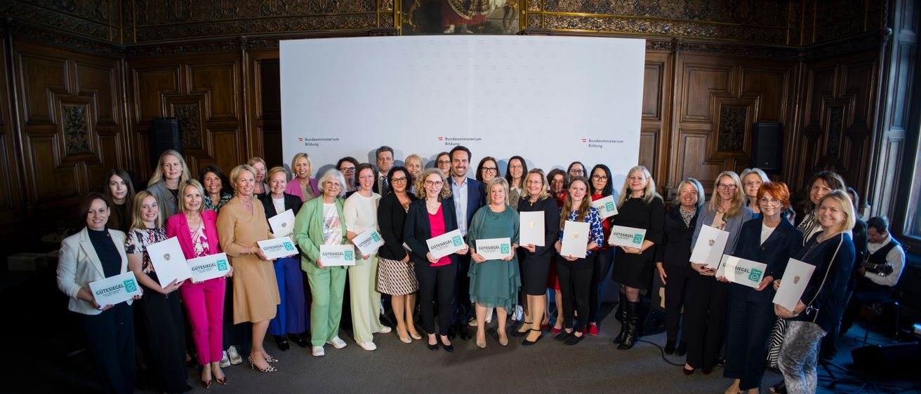 Eine Gruppe von Frauen und Männern in formeller Kleidung posiert mit Zertifikaten in der Hand für ein Foto. Sie stehen vor einem großen weißen Banner mit Logos und Text. Ein Gemälde einer historischen Figur ist hinter ihnen.