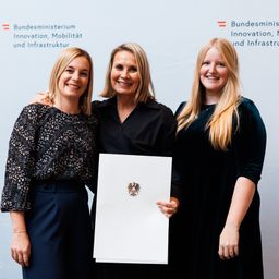 Drei Frauen stehen lächelnd vor einem weißen Hintergrund. Die mittlere Frau hält ein Zertifikat mit dem deutschen Wappen. Sie sind in formeller Kleidung gekleidet.