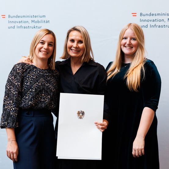 Drei Frauen stehen lächelnd vor einem weißen Hintergrund. Die mittlere Frau hält ein Zertifikat mit dem deutschen Wappen. Sie sind in formeller Kleidung gekleidet.