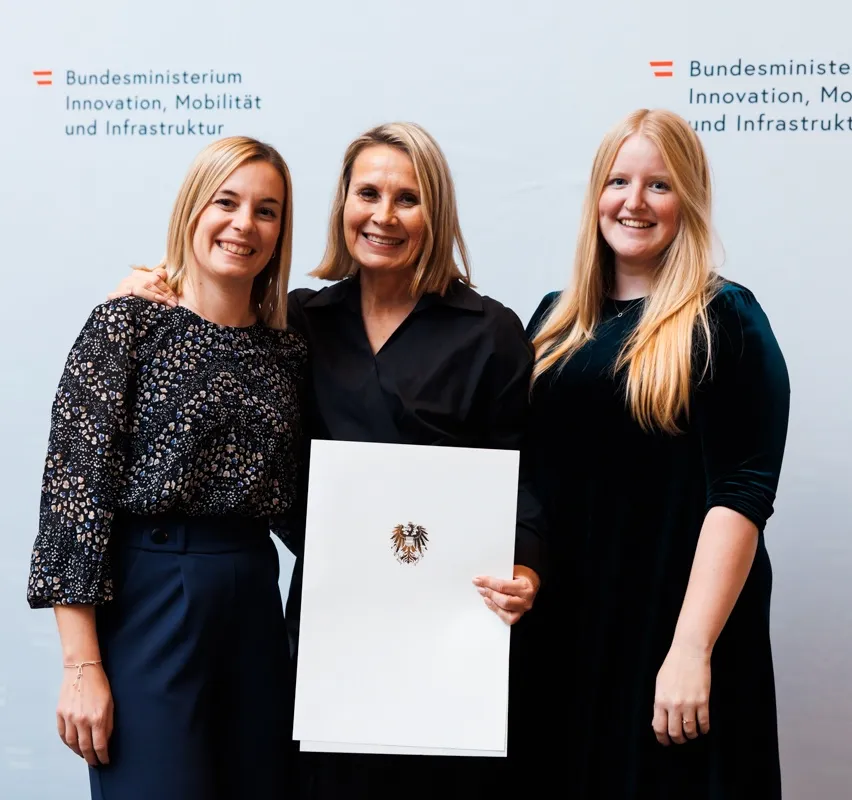 Drei Frauen stehen lächelnd vor einem weißen Hintergrund. Die mittlere Frau hält ein Zertifikat mit dem deutschen Wappen. Sie sind in formeller Kleidung gekleidet.