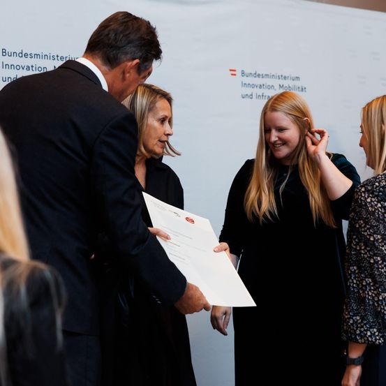 Ein Mann und zwei Frauen stehen vor einer weißen Wand. Der Mann überreicht einer der Frauen ein Zertifikat. Die Frau lächelt und schaut den Mann an.