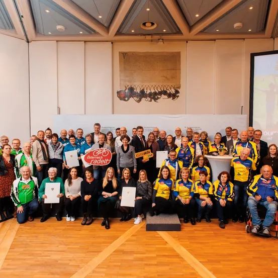 Eine Gruppe von Menschen, wahrscheinlich Radfahrer, posiert für ein Foto in einer Halle. Sie halten Urkunden in der Hand und stehen vor einem weißen Hintergrund mit einem Bild. Rechts ist eine Projektionsleinwand zu sehen.