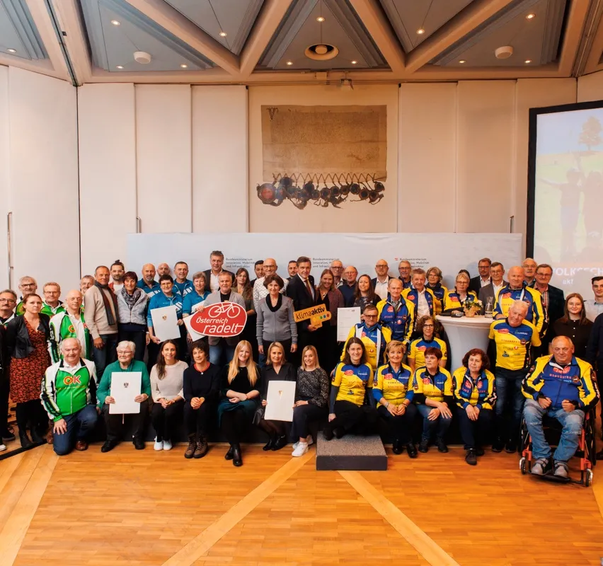 Eine Gruppe von Menschen, wahrscheinlich Radfahrer, posiert für ein Foto in einer Halle. Sie halten Urkunden in der Hand und stehen vor einem weißen Hintergrund mit einem Bild. Rechts ist eine Projektionsleinwand zu sehen.