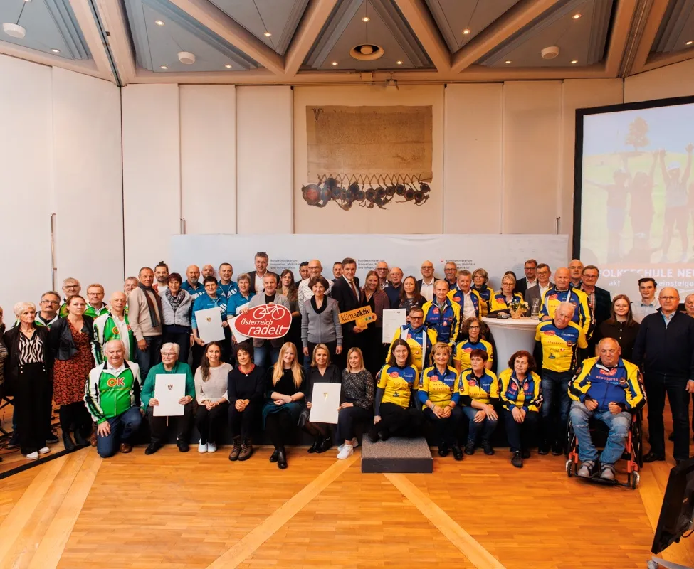 Eine Gruppe von Menschen, wahrscheinlich Radfahrer, posiert für ein Foto in einer Halle. Sie halten Urkunden in der Hand und stehen vor einem weißen Hintergrund mit einem Bild. Rechts ist eine Projektionsleinwand zu sehen.