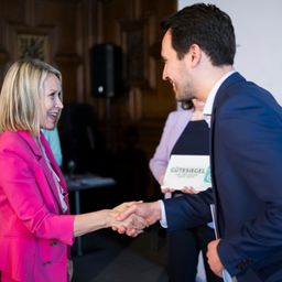 Eine Frau in einem rosa Blazer schüttelt einem Mann in einem blauen Anzug die Hand. Sie lächeln. Im Hintergrund hält eine Frau ein Zertifikat.