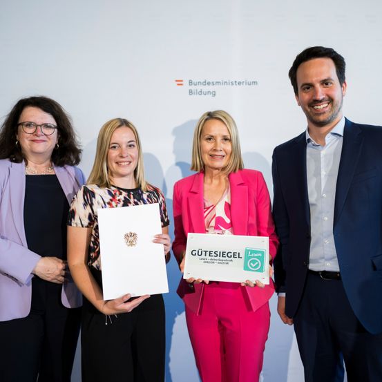 Vier Personen stehen zusammen vor einer weißen Wand. Zwei Frauen halten Urkunden, und die Frau in Pink trägt einen rosa Anzug. Der Mann auf der rechten Seite lächelt. Der Text 'Bundesministerium Bildung' ist auf der Wand sichtbar.