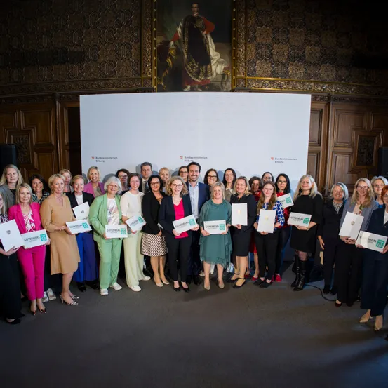 Eine Gruppe von Frauen und einem Mann posiert für ein Foto in einem Raum mit einer großen weißen Tafel und einem Porträt an der Wand. Sie halten alle Urkunden.