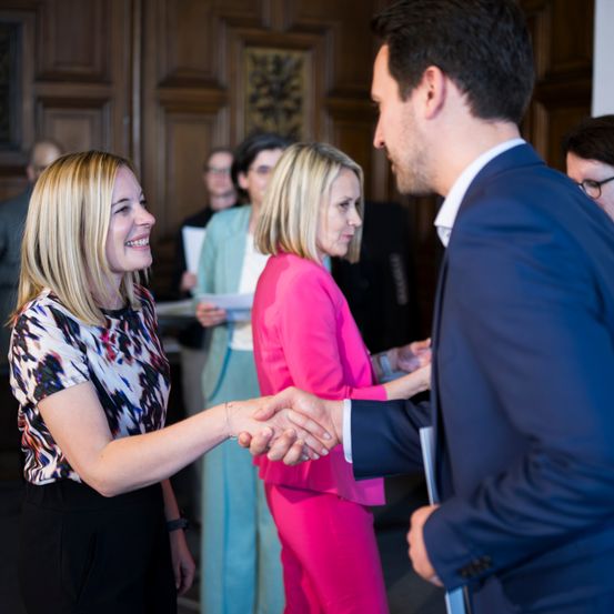 Ein Mann und eine Frau schütteln sich in einer professionellen Umgebung die Hände, umgeben von beobachtenden Kollegen. Die Frau lächelt, und eine andere Frau sieht ernst aus. Ein Holztür und ein Bilderrahmen sind im Hintergrund.