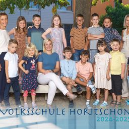 Eine Gruppe von Kindern und zwei Frauen posiert für ein Foto vor einem Gebäude, mit dem Text 'Volksschule Horitschon 2024-2025' unten.