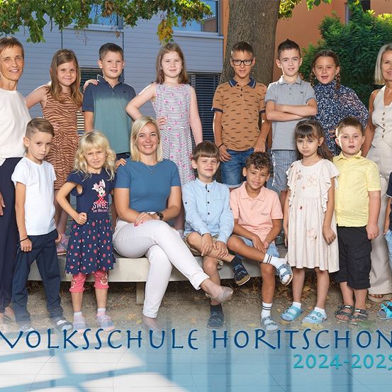 Eine Gruppe von Kindern und zwei Frauen posiert für ein Foto vor einem Gebäude, mit dem Text 'Volksschule Horitschon 2024-2025' unten.