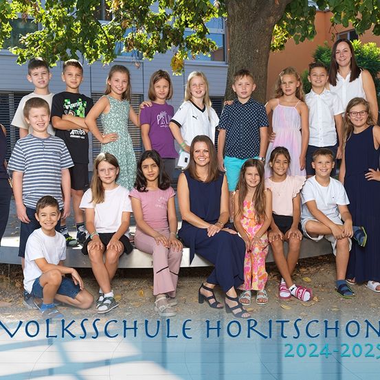 Eine Gruppe von Kindern und Erwachsenen posiert für ein Foto vor einem Baum, trägt Freizeitkleidung, Brillen und Turnschuhe. Das Bild enthält den Text 'VOLKSSCHULE HORITSCHOON 2024-2025'.