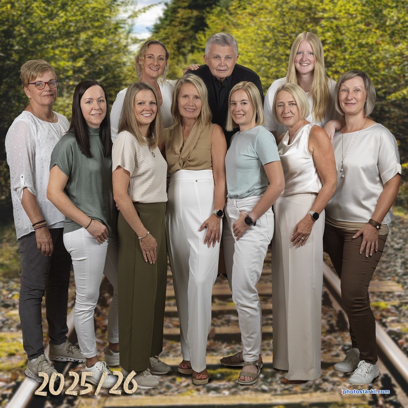 Gruppe von Menschen in übereinstimmenden weißen Hosen, die für ein Foto mit einem verblassten Hintergrund von Bäumen posieren. Das Bild hat einen Wasserzeichen mit dem Text 2025/26.