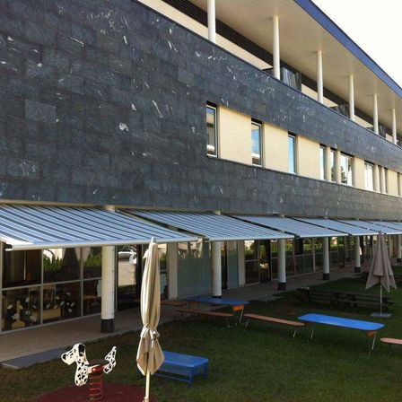 Ein modernes Gebäude mit schwarzer Fassade hat mehrere Fenster und Markisen, vor dem sich Picknicktische, Bänke und ein Spielplatz befinden.