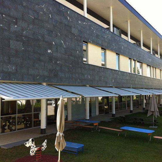 Ein modernes Gebäude mit schwarzer Fassade hat mehrere Fenster und Markisen, vor dem sich Picknicktische, Bänke und ein Spielplatz befinden.