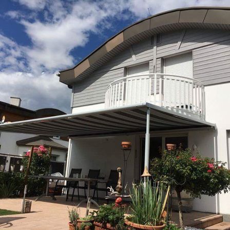Ein Haus mit einem Balkon, bedeckt von einem gestreiften Sonnendach, umgeben von Pflanzen, Stühlen und einem Tisch unter einem blauen Himmel mit Wolken.