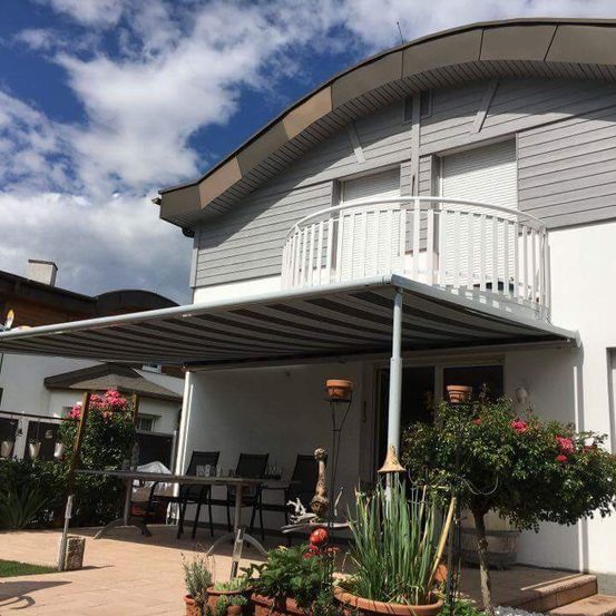 Ein Haus mit einem Balkon, bedeckt von einem gestreiften Sonnendach, umgeben von Pflanzen, Stühlen und einem Tisch unter einem blauen Himmel mit Wolken.
