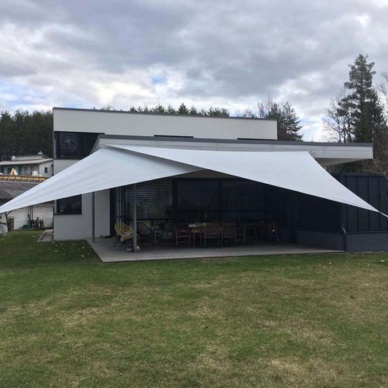 Ein modernes weißes Haus mit einem großen weißen Baldachin davor, umgeben von einem Rasen und Bäumen, mit einem Esstisch draußen aufgestellt.