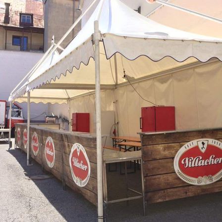 Eine Zeltbar mit einer hölzernen Theke, roten Kühlschränken und einem Logo. Sie befindet sich im Freien unter einem Zelt mit weißem Vordach.