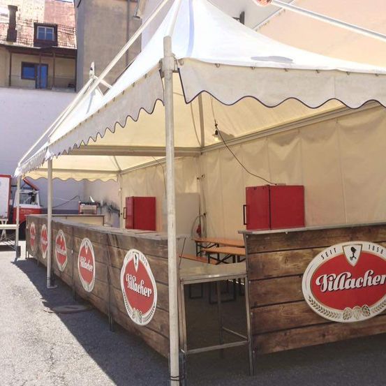 Eine Zeltbar mit einer hölzernen Theke, roten Kühlschränken und einem Logo. Sie befindet sich im Freien unter einem Zelt mit weißem Vordach.