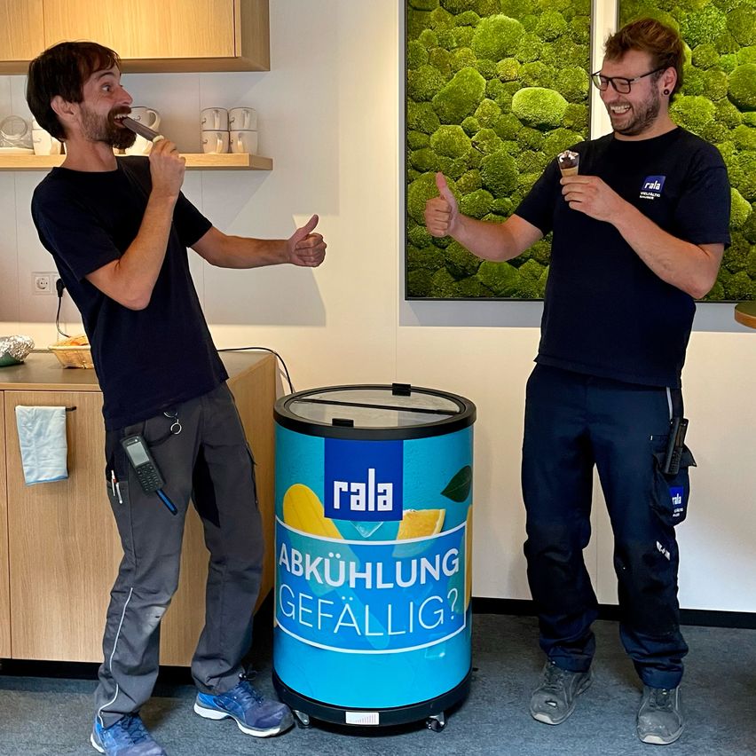 Zwei Männer stehen neben einem blauen Kühlschrank mit der Aufschrift 'Abkühlung Gefällig?'. Ein Mann gibt ein Daumen hoch, während er einen Eisbecher isst, und der andere hält einen Eisbecher in der Hand.