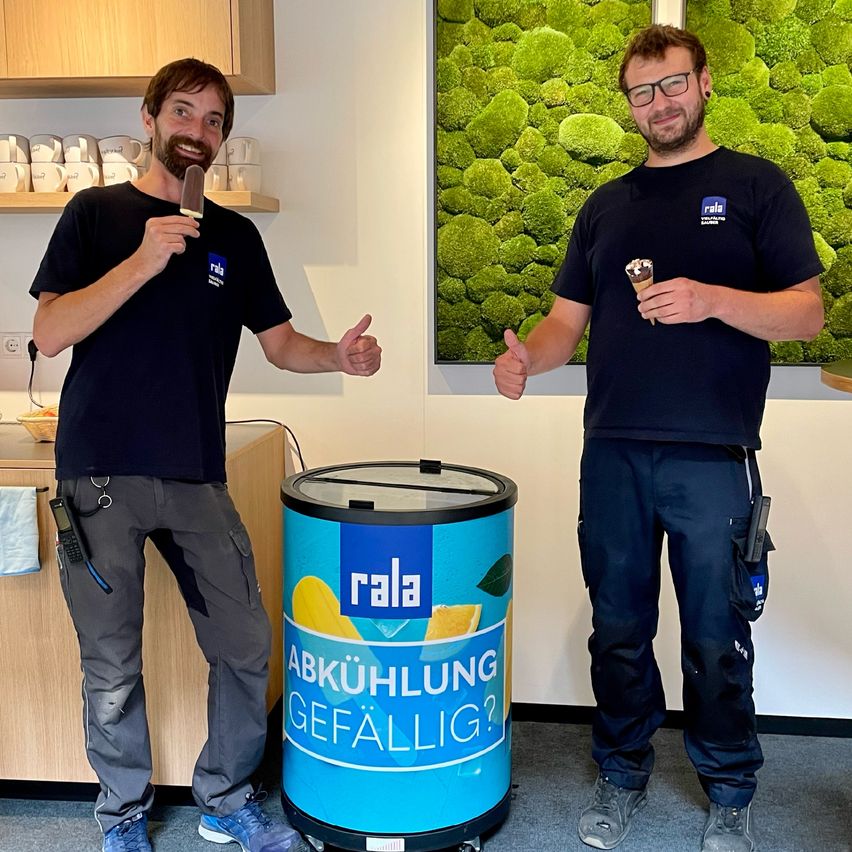 Zwei Männer stehen neben einem blauen Fass mit der Aufschrift 'Abkühlerung Gefällig?'. Sie halten Eiswaffeln in der Hand und geben Daumen hoch. Hinter ihnen befindet sich eine grüne Mooswand und ein Holzschrank mit Tassen.